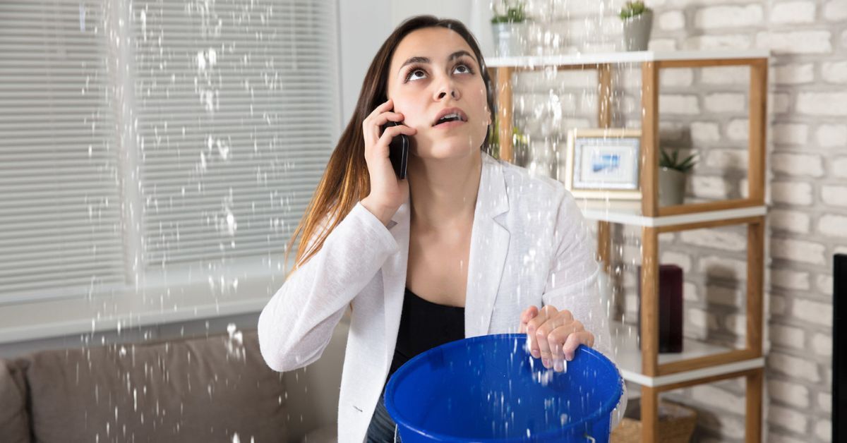 Woman on phone while holding a bucket to catch water dripping down from a leak.